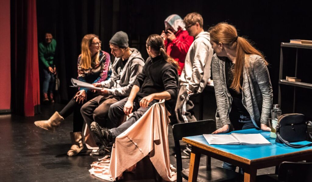 Acting students rehearsing together in a classroom setting, sitting and reading scripts while receiving guidance from their tutor.